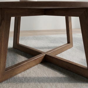 May include: A low-angle view of a wooden coffee table with a geometric base. The table has a dark brown finish and a square top. The base features an X-shaped design, with the legs intersecting in the center. A vase with branches is on top.