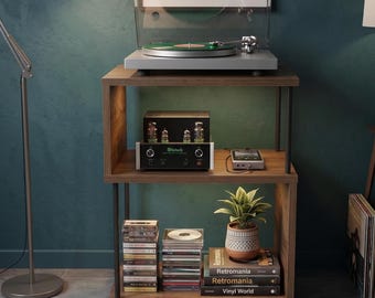 May include: A wooden shelving unit with a turntable, amplifier, CDs, and books. A framed geometric art print hangs above the turntable. A small potted plant and a floor lamp are also visible.