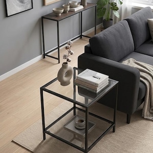 May include: A square end table with a black metal frame and two clear glass shelves. The top shelf holds a beige ceramic vase with cotton stems and a stack of three books. The bottom shelf holds a small square tray with a white ceramic cup.