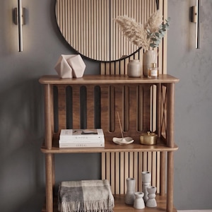May include: A wooden bookshelf with three tiers and rounded corner posts. The top shelf holds a geometric sculpture and a vase with dried plants. The middle shelf contains two books labeled Kinfolk and Cereal, a small dish, and a gold container. A folded plaid blanket and three gray vases sit on the bottom shelf.