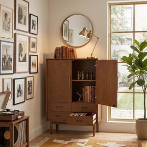 Op de afbeelding: Een houten kast in mid-century modern stijl met laden en deuren, waarvan &eacute;&eacute;n open is. Een goudkleurige bureaulamp en een ronde spiegel staan erop. Een verzameling ingelijste kunst en een platenspeler zijn ook zichtbaar.