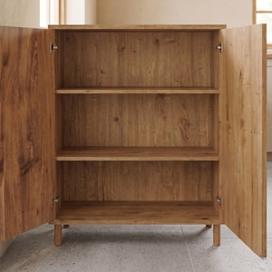 Puede incluir: Un mueble de madera marr&oacute;n claro con dos puertas abiertas, que revelan tres estantes. El mueble es de madera con veta natural y se apoya sobre patas cortas y redondeadas. Las puertas est&aacute;n articuladas y se abren hacia los lados.