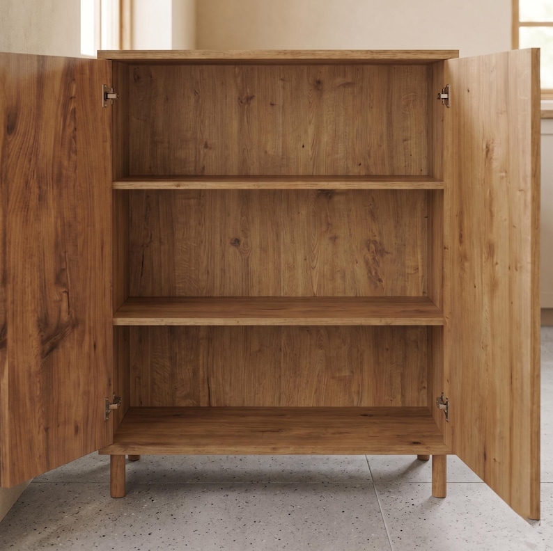 Puede incluir: Un mueble de madera marr&oacute;n claro con dos puertas abiertas, que revelan tres estantes. El mueble es de madera con veta natural y se apoya sobre patas cortas y redondeadas. Las puertas est&aacute;n articuladas y se abren hacia los lados.