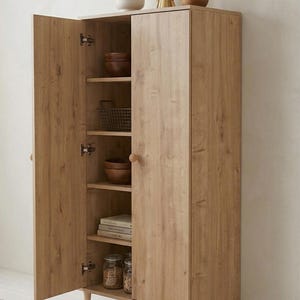 May include: A light-colored wooden cabinet with two doors, one open to reveal shelves holding bowls, a wire basket, books, and jars. A small plant in a white pot and decorative items sit on top. The cabinet is on short legs.