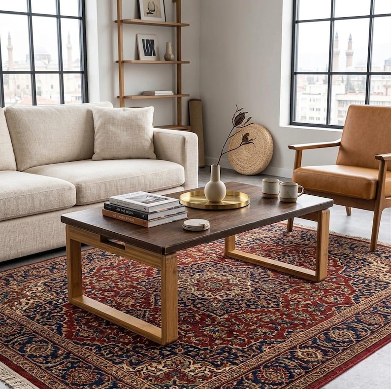 Op de afbeelding: Een woonkamer met een beige bank, een bruine houten salontafel en een bruine leren fauteuil. De salontafel heeft boeken, een gouden dienblad en mokken. Een patroonkleed in rood, blauw en beige bedekt de vloer.