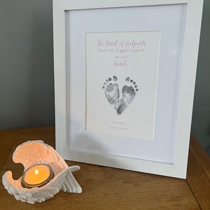 May include: A white framed print featuring baby footprints and the text "the tiniest of footprints leave the biggest impact on our hearts." An angel wing candle holder with a lit candle is in front of the frame.