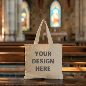 May include: A white canvas tote bag with the text "YOUR DESIGN HERE" printed in gray on the front. The bag is sitting on a wooden bench in a church.