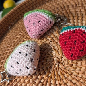 May include: Three handmade crochet watermelon keychains. One is a pink slice with green rind, another is a red heart-shaped watermelon with a green rind, and the third is a white oval with a green rind. All have black seed details.
