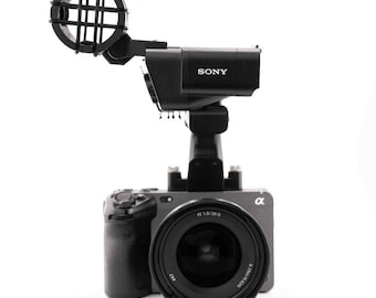 May include: A black Sony camera with a lens attached and a microphone on top. The camera has a black and silver body and the lens says "FE 1.8/20 G".