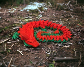 May include: A red and green origami dragon made of paper, lying on the ground in a forest setting.