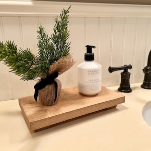 May include: A wooden tray with a small potted green plant and a bottle of goat milk hand soap labelled "Crabtree & Evelyn" on a white countertop.