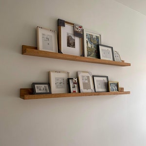 May include: Two wooden floating shelves on a white wall, displaying framed pictures. The frames vary in colour, including black, white, and gold. The shelves are made of light-coloured wood. The pictures are of different sizes.