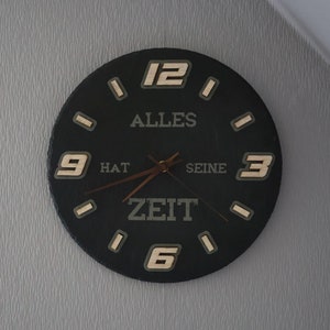 May include: A circular black wall clock with wooden numerals and hands. The clock face displays the German text "ALLES HAT SEINE ZEIT" in a light colour. The numbers 12, 3, 6, and 9 are clearly visible.