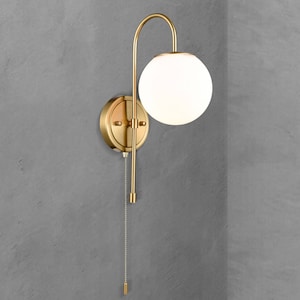 Könnte beinhalten: Eine goldene Wandleuchte mit einem weißen Globus-Schirm und einer Zugkette. Die Leuchte hat einen gebogenen Arm und einen runden Sockel.
