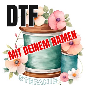 Puede incluir: Una bobina de hilo de color turquesa y marrón con el texto "DTF MIT DEINEM NAMEN" en letras rojas y el nombre "STEFANIE" en letras azules. La bobina está rodeada de flores rosas y blancas.