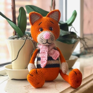 Puede incluir: Un juguete de gato de crochet naranja con rayas negras, que lleva una bufanda rosa. El gato tiene una nariz rosa y ojos negros.