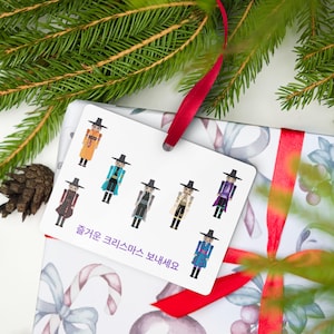 May include: A gift tag with illustrations of figures in traditional Korean attire, in various colors, is attached to a wrapped gift. The gift is wrapped in patterned paper and tied with a red ribbon. Pine needles and a pine cone are in the background.
