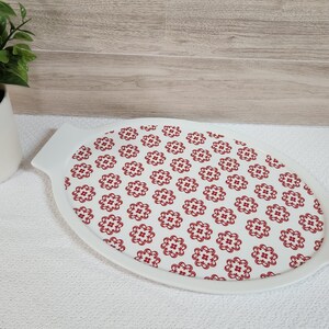 May include: Oval white serving dish with a red floral pattern on a white background. The dish has handles on each end. A small potted plant is in the background.