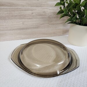 May include: A vintage, brown glass casserole dish lid. The lid is oval-shaped with a rounded handle and a slightly raised edge. The glass has a smoky, transparent appearance. The lid is resting on a white surface, with a plant in a white pot in the background.