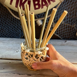 Può includere: Un insieme di cannucce di bambù in un barattolo di vetro decorativo. Il barattolo è avvolto in vimini intrecciato e adornato con conchiglie. Le cannucce sono in varie tonalità di beige e marrone chiaro, un prodotto ecologico.