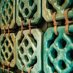 May include: A close-up shot of several jade-green decorative tiles with detailed openwork designs. Each rectangular tile, with rounded corners, is linked by brown rope. The tiles, with a glossy finish, are arranged in a grid pattern.