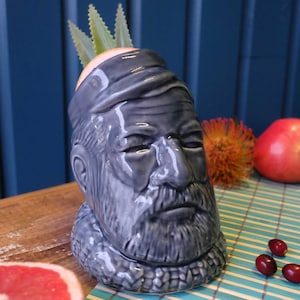 Puede incluir: Un mug tiki de cerámica gris con forma de cabeza de hombre con barba. El mug tiene una planta verde que crece en la parte superior.