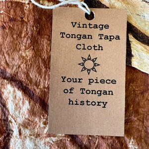 May include: A rectangular tan paper tag with a white string attached, featuring black text that reads Vintage Tongan Tapa Cloth, a sun icon, and the text Your piece of Tongan history, resting on a textured brown and cream patterned fabric.