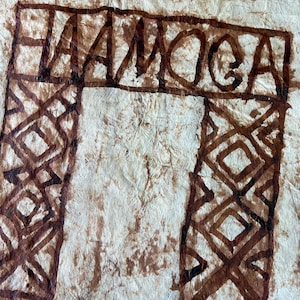 May include: A textured, off-white fibrous material featuring a dark brown hand-painted design. The artwork includes a rectangular border at the top containing the text "AAMOCA" and two vertical columns decorated with geometric diamond patterns.