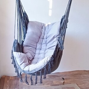Puede incluir: Una silla colgante gris con un cojín gris y blanco. La silla tiene flecos alrededor de los bordes y está hecha de cuerda.