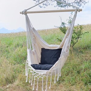 Puede incluir: Una silla hamaca blanca con un cojín negro, colgada de una rama de árbol en un campo de hierba.