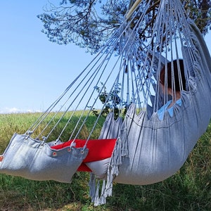May include: A person relaxing in a gray hammock with fringe details. The hammock is hanging from a tree branch and the person is wearing red pants and white socks.