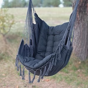 Puede incluir: Una silla hamaca negra con detalles de flecos cuelga de una rama de árbol. La silla tiene un cojín grueso y acolchado y está hecha de una tela suave y tejida.