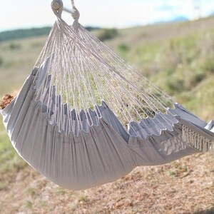 May include: A gray hammock with fringe hanging from the bottom. The hammock is suspended between two trees in a grassy field.