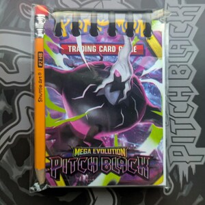 Könnte beinhalten: Ein kleines Notizbuch mit Spiralbindung und einem Pokemon Pitch Black Mega Evolution Motiv auf dem Einband, begleitet von einem gelben Shuttle Art #2 HB Bleistift. Das Notizbuch ist in transparenter Folie verpackt und zeigt den Text Pokemon Trading Card Game.
