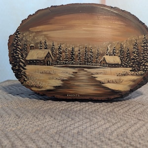 May include: A hand-painted wooden slice with a winter scene depicting two small houses with snow-covered roofs, a winding path, and a line of trees in the background. The wood is a dark brown color and the painting is done in shades of brown, white, and black.