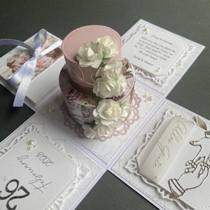 May include: A handcrafted paper gift box with a small, round, pink and floral-patterned box decorated with white paper flowers. The box is on a white, lace-edged card with text and pearls. A pale blue ribbon is visible.
