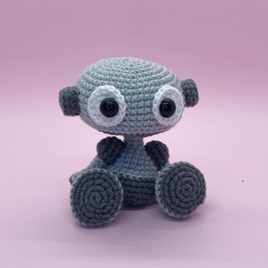 Könnte beinhalten: Ein graues Häkelroboter-Spielzeug mit großen weißen Augen und schwarzen Pupillen. Der Roboter sitzt mit gekreuzten Beinen und hat einen kleinen, runden Körper.