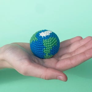 May include: A crocheted globe, featuring blue oceans, green continents, and a white polar region. The handmade sphere is held in a hand against a light green background. The globe is a representation of the Earth.