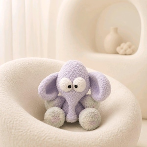 May include: A light purple crocheted elephant plush toy with a textured, fuzzy yarn finish. The elephant features large, round white eyes with black pupils, prominent ears, and a central trunk. The lower limbs are a lighter, mottled pastel shade.