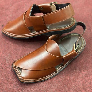 K&ouml;nnte beinhalten: Ein Paar braune Ledersandalen mit Kn&ouml;chelriemen und Schnallenverschluss. Die Sandalen haben eine gen&auml;hte Sohle und ein Kreuzmuster &uuml;ber dem Fu&szlig;.