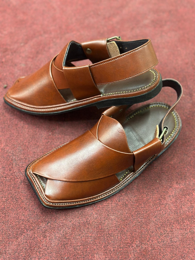 K&ouml;nnte beinhalten: Ein Paar braune Ledersandalen mit Kn&ouml;chelriemen und Schnallenverschluss. Die Sandalen haben eine gen&auml;hte Sohle und ein Kreuzmuster &uuml;ber dem Fu&szlig;.