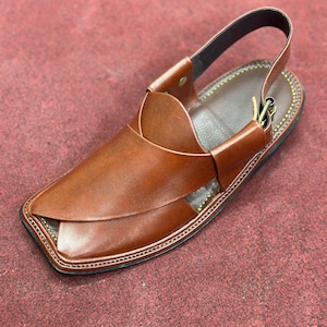 K&ouml;nnte beinhalten: Braune Ledersandale mit Riemen. Die Sandale hat eine quadratische Zehenpartie und einen gen&auml;hten Rand. Der obere Teil der Sandale hat ein einzigartiges Design mit &uuml;berlappenden Lederst&uuml;cken. Die Sandale hat eine dunkelbraune Sohle und einen schwarzen Riemen.