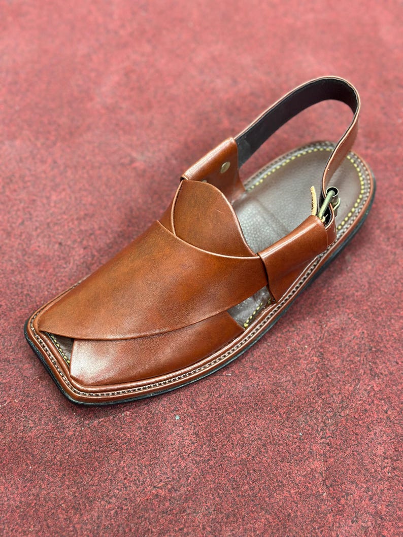 K&ouml;nnte beinhalten: Braune Ledersandale mit Riemen. Die Sandale hat eine quadratische Zehenpartie und einen gen&auml;hten Rand. Der obere Teil der Sandale hat ein einzigartiges Design mit &uuml;berlappenden Lederst&uuml;cken. Die Sandale hat eine dunkelbraune Sohle und einen schwarzen Riemen.