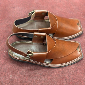 K&ouml;nnte beinhalten: Ein Paar braune Ledersandalen mit quadratischer Zehenpartie. Die Sandalen haben einen Kn&ouml;chelriemen und Kontrastn&auml;hte. Das Schuhwerk wird auf einer roten Oberfl&auml;che pr&auml;sentiert.