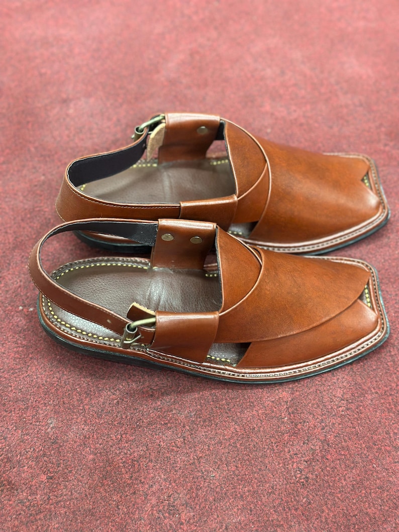 K&ouml;nnte beinhalten: Ein Paar braune Ledersandalen mit quadratischer Zehenpartie. Die Sandalen haben einen Kn&ouml;chelriemen und Kontrastn&auml;hte. Das Schuhwerk wird auf einer roten Oberfl&auml;che pr&auml;sentiert.