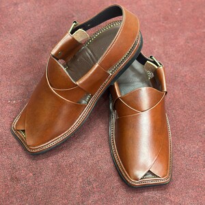 K&ouml;nnte beinhalten: Ein Paar braune Peshawari-Sandalen aus Leder. Die Sandalen haben ein unverwechselbares Design mit einem gekreuzten Riemen &uuml;ber dem Oberteil und einem Kn&ouml;chelriemen. Die Kanten sind mit einem Kontrastfaden gen&auml;ht, und die Sohlen sind schwarz.