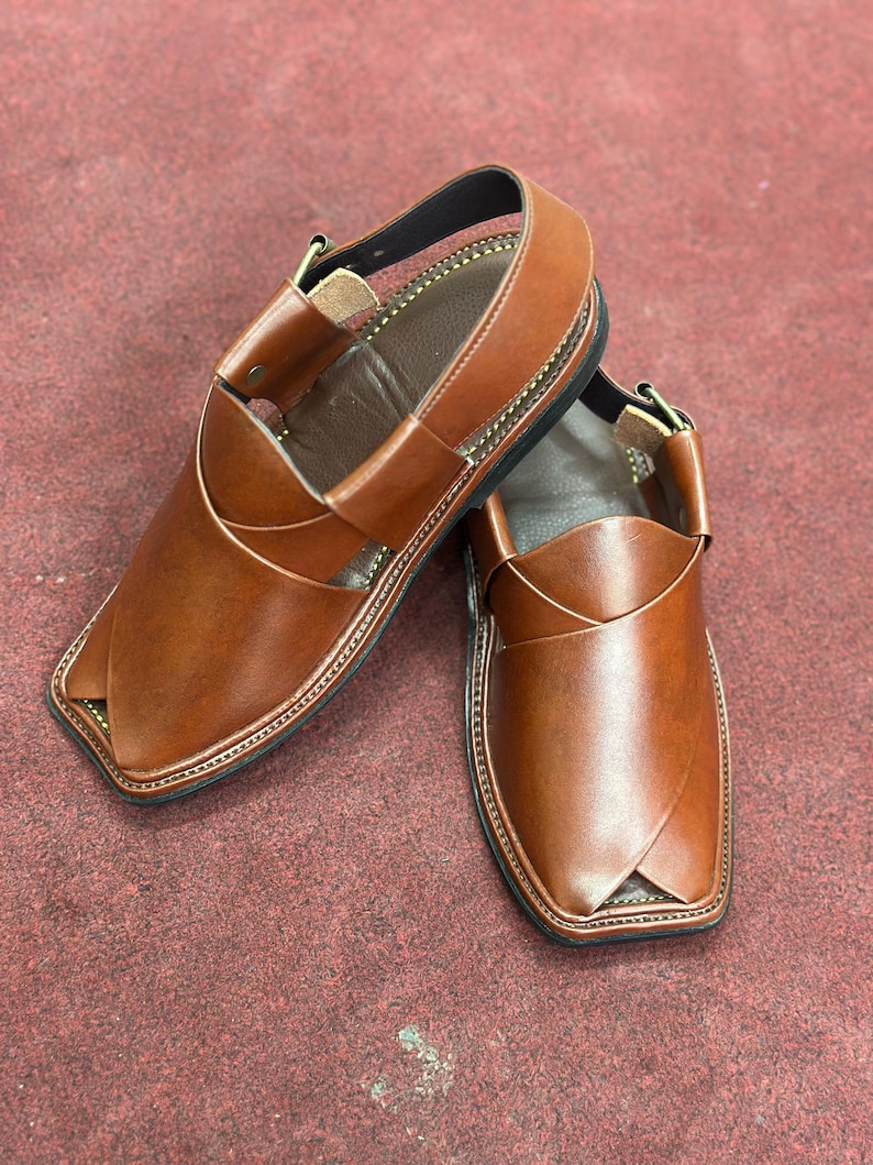 K&ouml;nnte beinhalten: Ein Paar braune Peshawari-Sandalen aus Leder. Die Sandalen haben ein unverwechselbares Design mit einem gekreuzten Riemen &uuml;ber dem Oberteil und einem Kn&ouml;chelriemen. Die Kanten sind mit einem Kontrastfaden gen&auml;ht, und die Sohlen sind schwarz.