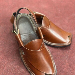 K&ouml;nnte beinhalten: Ein Paar braune Peshawari-Sandalen aus Leder. Die Sandalen haben eine quadratische Zehenpartie mit einem Kn&ouml;chelriemen. Die Kanten sind mit einem Kontrastfaden gen&auml;ht, und die Sohle ist schwarz.