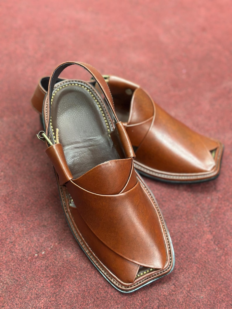 K&ouml;nnte beinhalten: Ein Paar braune Peshawari-Sandalen aus Leder. Die Sandalen haben eine quadratische Zehenpartie mit einem Kn&ouml;chelriemen. Die Kanten sind mit einem Kontrastfaden gen&auml;ht, und die Sohle ist schwarz.