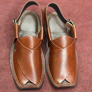 K&ouml;nnte beinhalten: Ein Paar braune Ledersandalen. Die Sandalen haben ein unverwechselbares Design mit einem Riemen um den Kn&ouml;chel und einer quadratischen Zehenpartie. Das Leder hat eine gl&auml;nzende Oberfl&auml;che. Die Sohlen sind schwarz.
